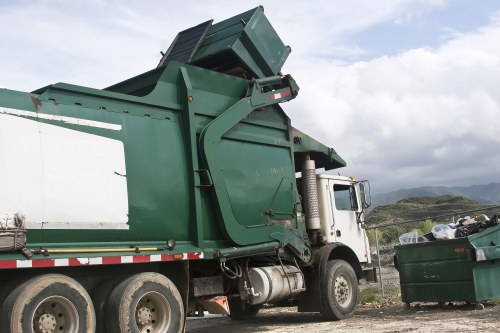 A large green skip mounted on a white and green waste collection truck with a hydraulic lifting mechanism in a rural outdoor setting, positioned on a gravel surface. The skip's sides are made of metal with a slightly textured, painted finish, and it is partially open at the top, revealing some mixed waste inside. The truck features a white cab with visible side mirrors and exhaust pipes, alongside a series of black and metallic wheels, some of which are in the foreground. In the background, there is a countryside landscape with rolling hills, rocky terrain, and partly cloudy sky, suggesting a disposal or rubbish removal operation typical of services offered by Garden Clearance Chingford, serving areas around Epping Forest, Chingford, and nearby postcode districts.