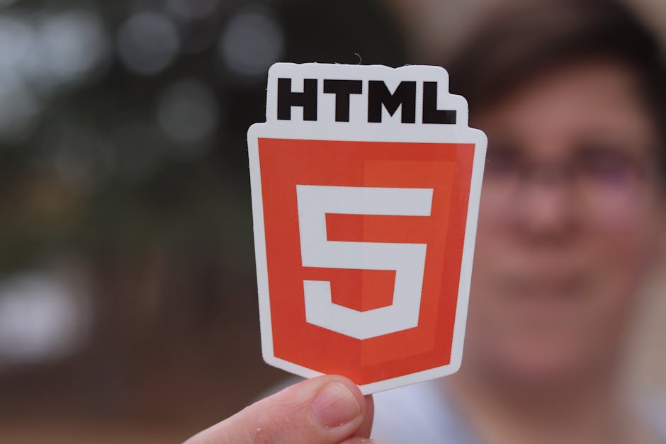 A person outdoors is holding a rectangular sticker featuring the HTML5 logo, which consists of a bold white number '5' over an orange shield-shaped background with the word 'HTML' printed above in bla