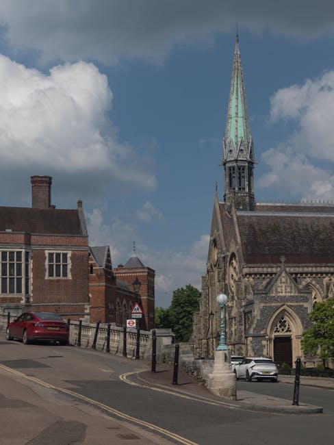 A street scene featuring a historic church with a tall, pointed spire covered in aged patina copper, situated to the right of the image. The church's stone façade displays intricate Gothic architectur